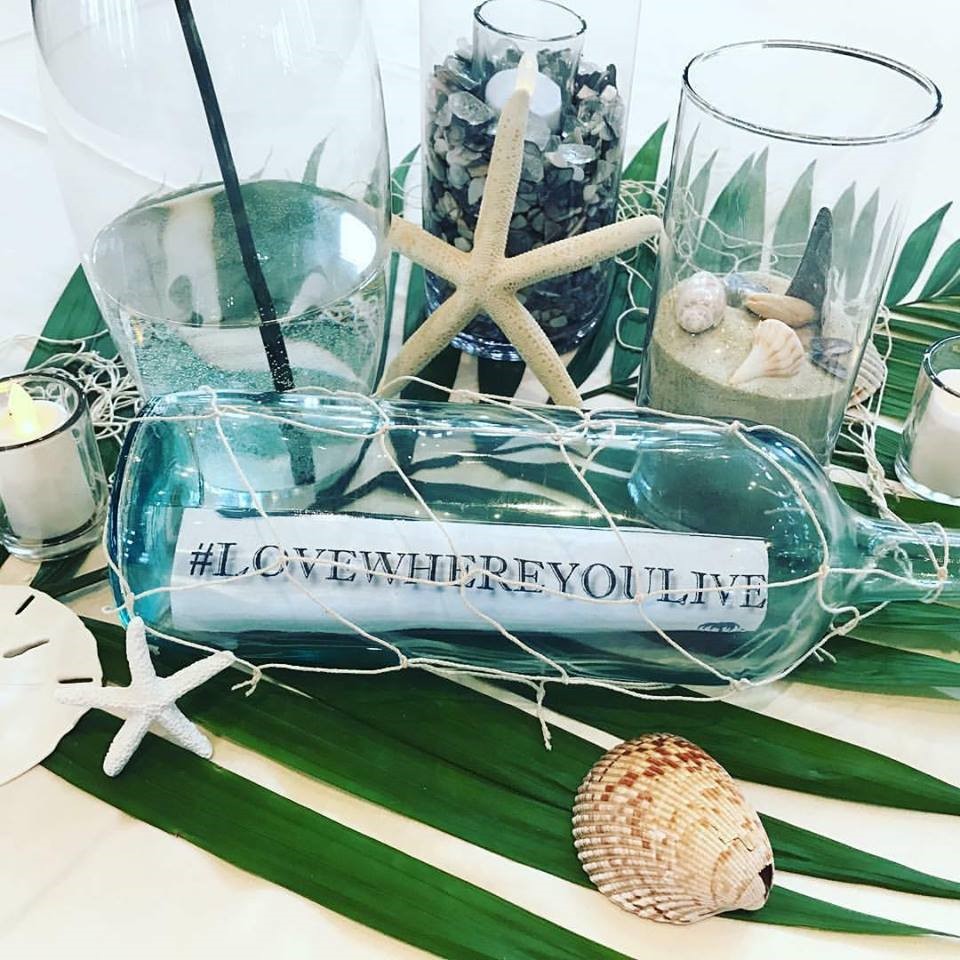 tropical table decoration with a bottle displaying message that says # Love Where You Live, with sand, shells, rocks, and artificial candles.